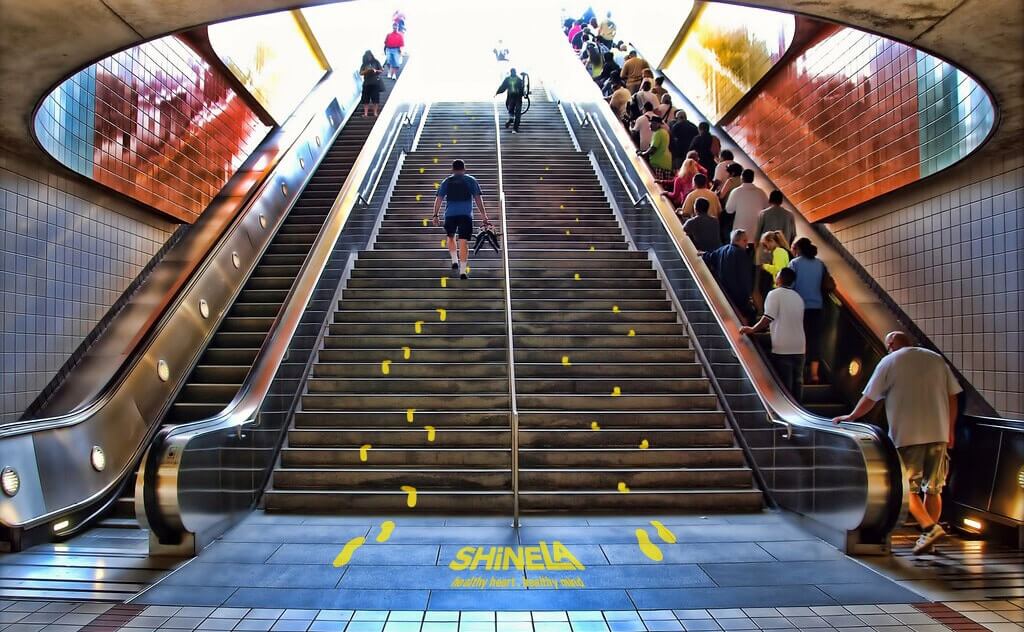 Photo from the bottom of a staircase/escalator combo in the metro, the stairs have a mockup encouraging people to stay fit with call to action footprints